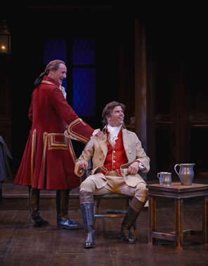 Archer in long red coat with gold embroidery and riding boots, his hair in a pony tail, leans on the chair in which sits Aimwell, in tan coat and britches, red vest, and fluffy white shirt, black riding boots with brown tops, and long hair. Next to him is a table with pewter ewer and tankards